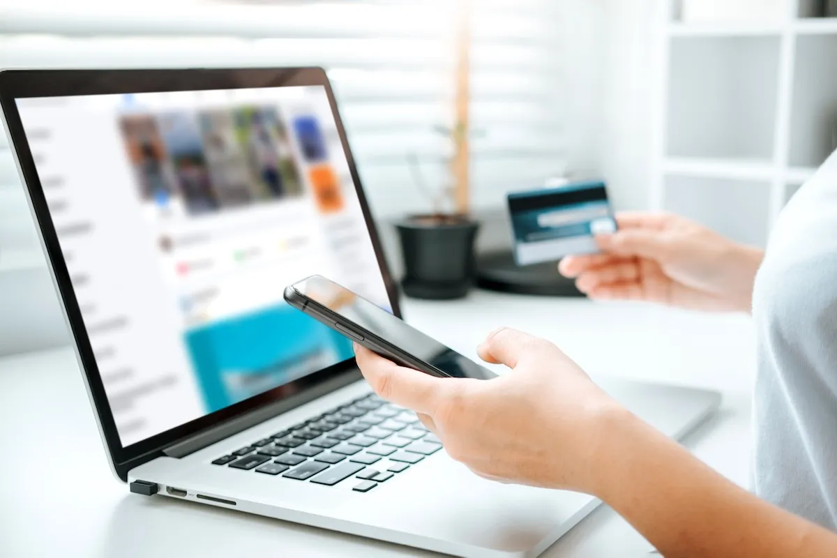 Person holding a smartphone and credit card while using a laptop on a desk, possibly browsing websites for bookkeepers, suggesting online shopping or digital banking activity.