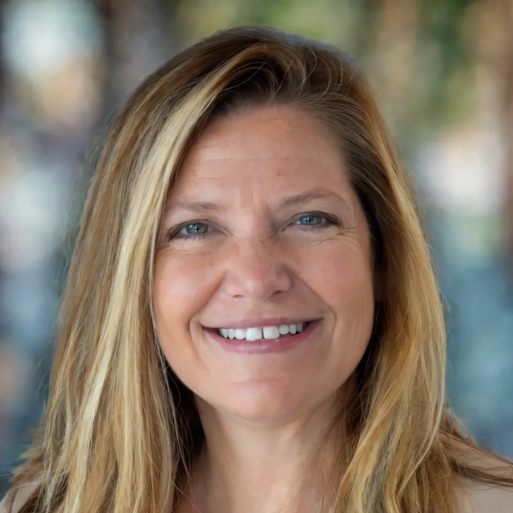 A woman with long blonde hair smiles at the camera against a blurred outdoor background, perfect for websites for bookkeepers seeking a professional yet approachable image.