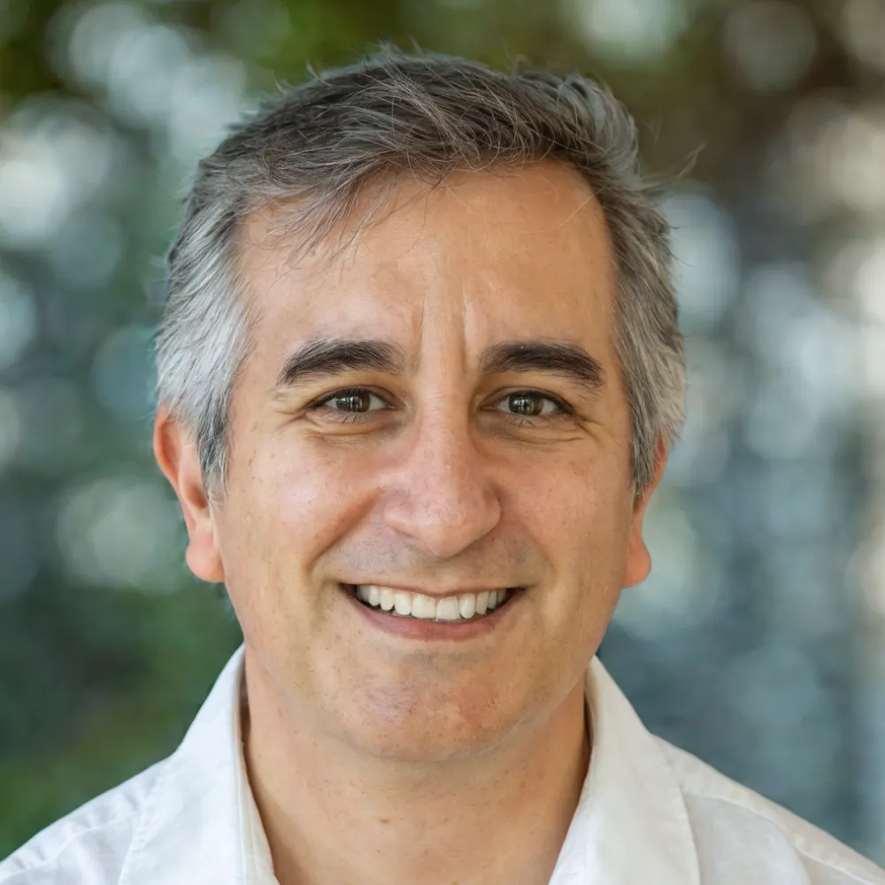 A middle-aged man with gray hair smiles at the camera, wearing a white shirt, with a blurred green and blue background—perfect for websites for bookkeepers seeking a professional, approachable image.