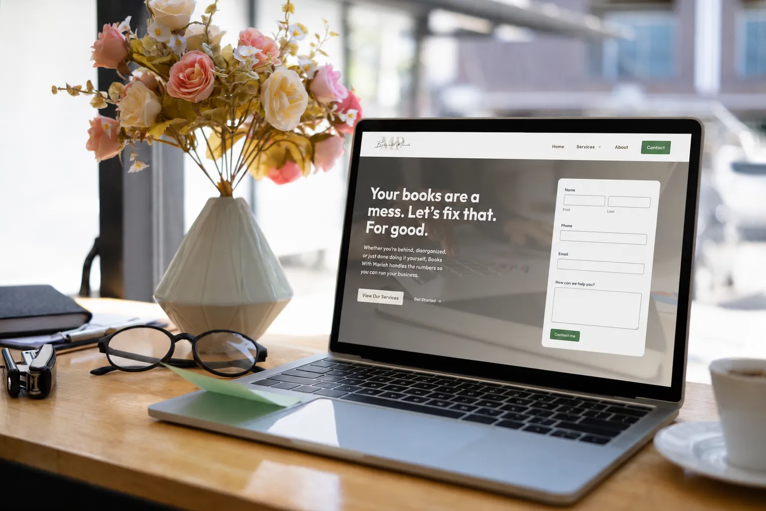 A laptop on a wooden desk displays a bookkeeping website. Nearby are a vase of flowers, a notebook, eyeglasses, and a pen. Sunlight comes through a window in the background.